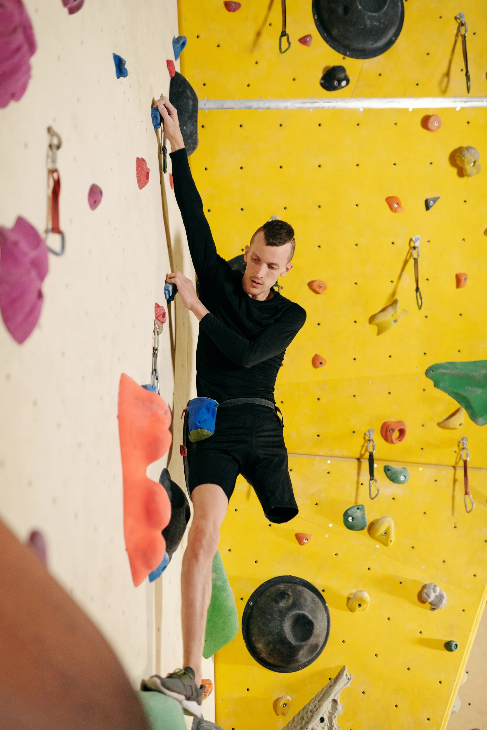 A climber in a black long-sleeve shirt ascends a vibrant yellow climbing wall, navigating colorful holds of various shapes.