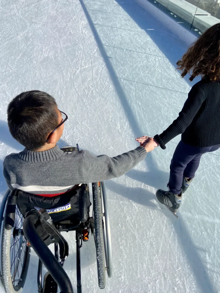 A child in a wheelchair holds hands with a friend while ice skating, enjoying the moment on a sunlit ice rink.
