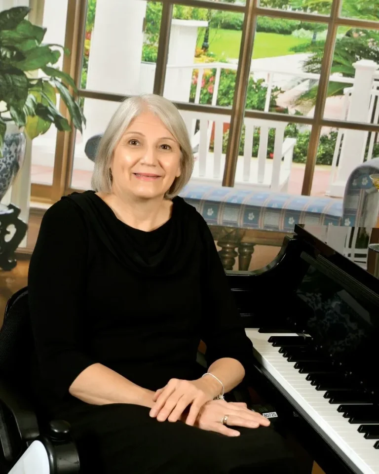A woman in a black dress sits next to a piano, with a lush garden visible through large windows behind her.