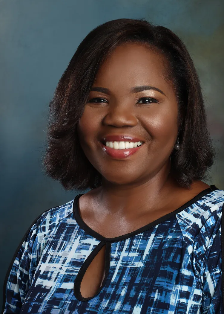 A woman with dark, wavy hair wears a blue patterned shirt, posing in front of a soft, muted background.
