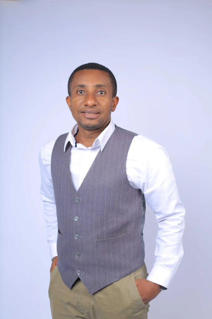 A man in a gray pinstriped vest over a white shirt poses confidently with his hands in his pockets against a light background.