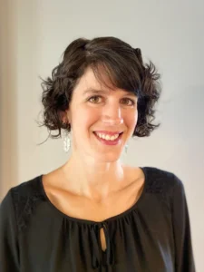 A woman with short, curly hair wearing a black blouse with a tie detail, standing against a light-colored wall.