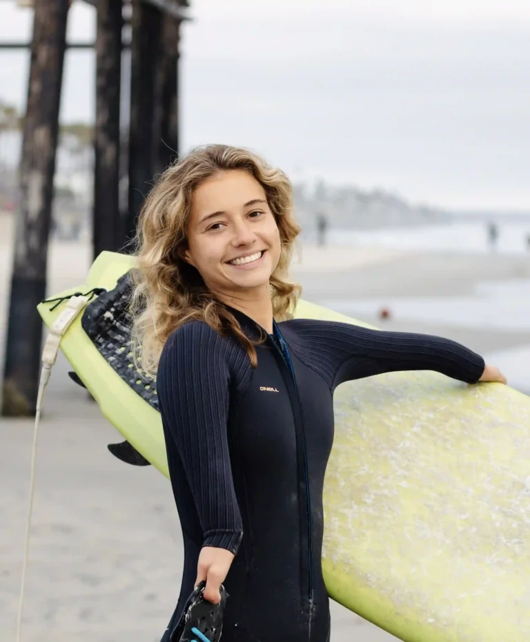 A person in a black wetsuit stands on a beach, holding a yellow surfboard, with ocean waves and a cloudy sky in the background.