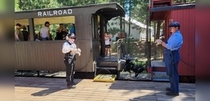 Two conductors in vintage attire interact near a train car, with a young child peeking out from inside, surrounded by trees.
