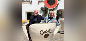 A couple sits inside a giant chocolate cup with a heart-shaped sign, enjoying a sunny day in a quaint outdoor setting.