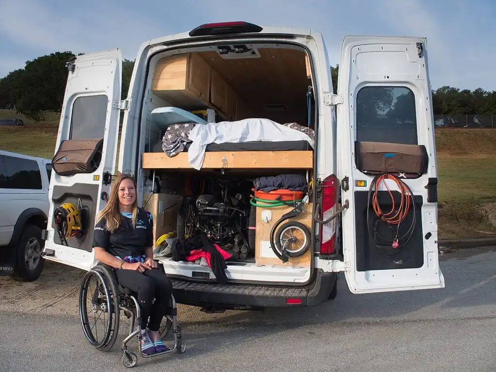 A woman in a wheelchair sits beside an open van filled with organized gear, a bed, and outdoor equipment, set against a grassy background.
