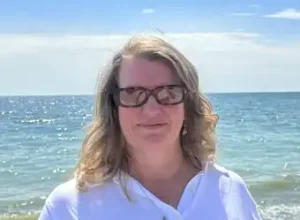 A person stands by the shore, wearing a white shirt, with the ocean and a bright sky in the background.