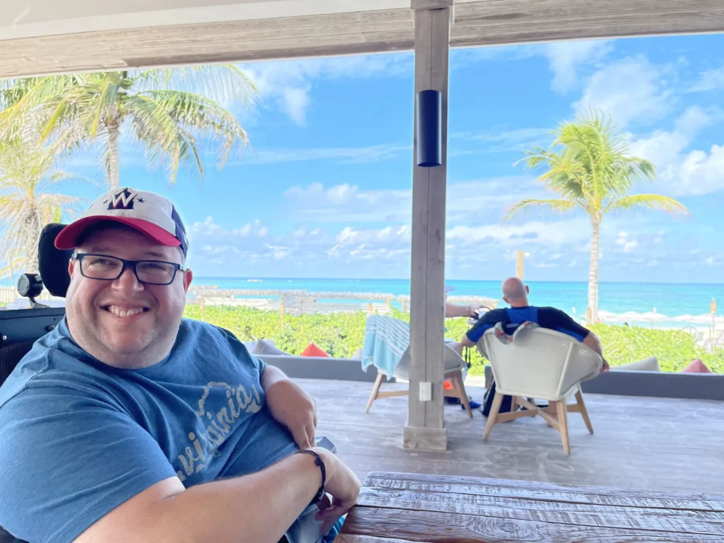 View of a tropical beach with palm trees and turquoise water, seen from a cozy outdoor seating area. Bright blue sky with fluffy clouds.