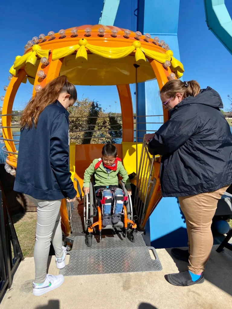 A child in a wheelchair prepares to board a brightly colored amusement ride, assisted by two adults on a sunny day.