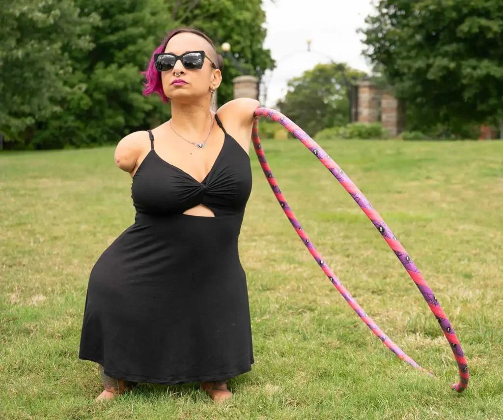 A woman in a black dress poses confidently on grass, holding a colorful hula hoop, showcasing her vibrant personality and strength.