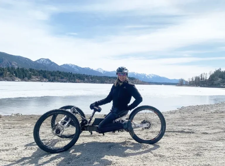 A person sits on a handbike on a sandy beach next to a frozen lake, with mountains in the background under a bright sky.