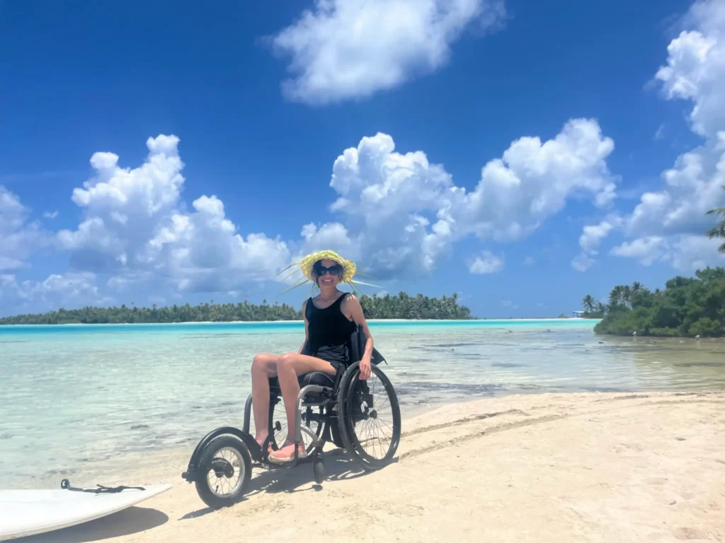 A person in a wheelchair wearing a sunhat enjoys a sunny beach scene with turquoise water and lush palm trees in the background.