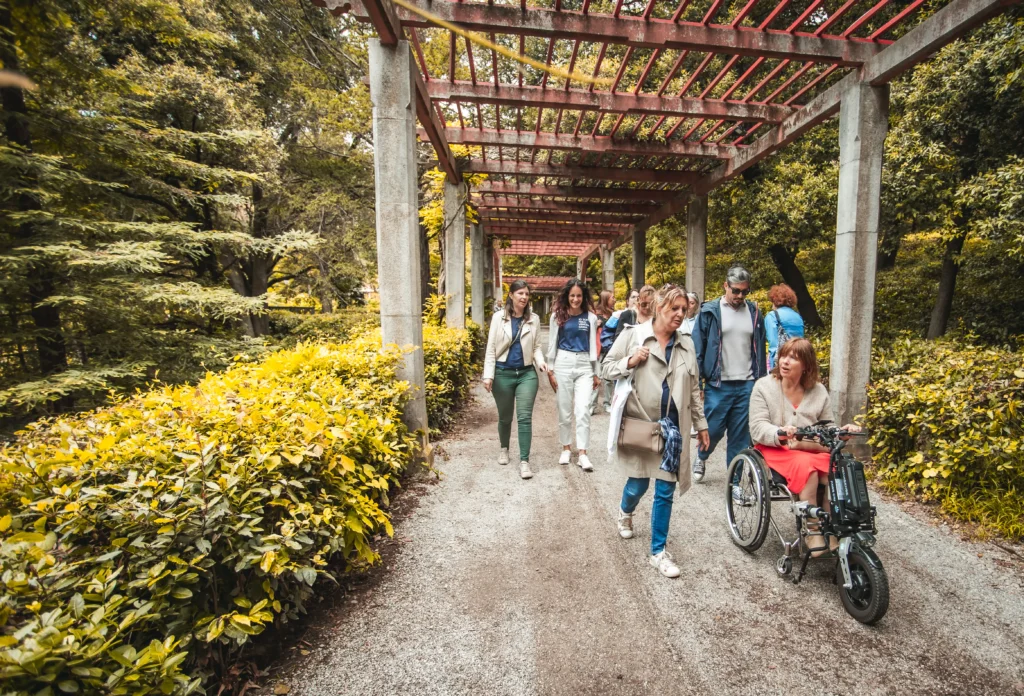 A diverse group of people strolls along a garden path, accompanied by a wheelchair user, under a trellis surrounded by greenery.