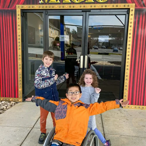 Three children pose excitedly in front of a vibrant entrance adorned with "Magic" signage, ready for an adventure.