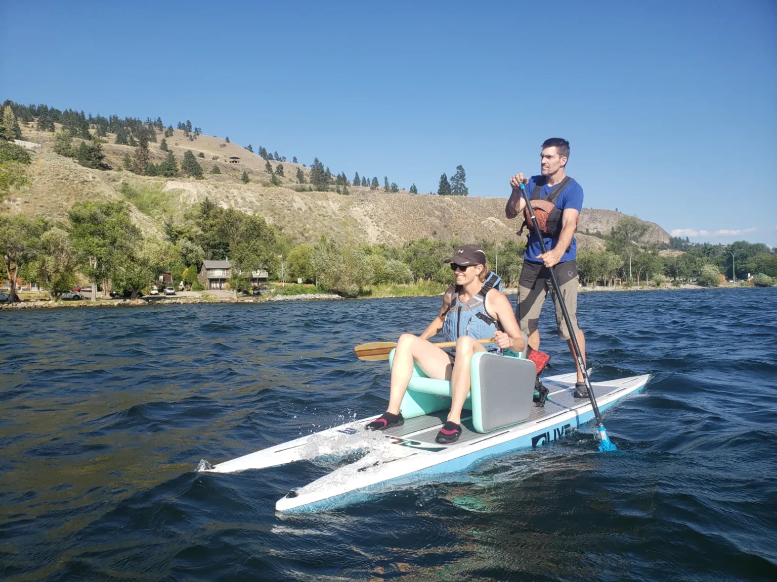 Couple on a stand-up paddleboard with a seated passenger, enjoying a sunny day on a lake with hills in the background.