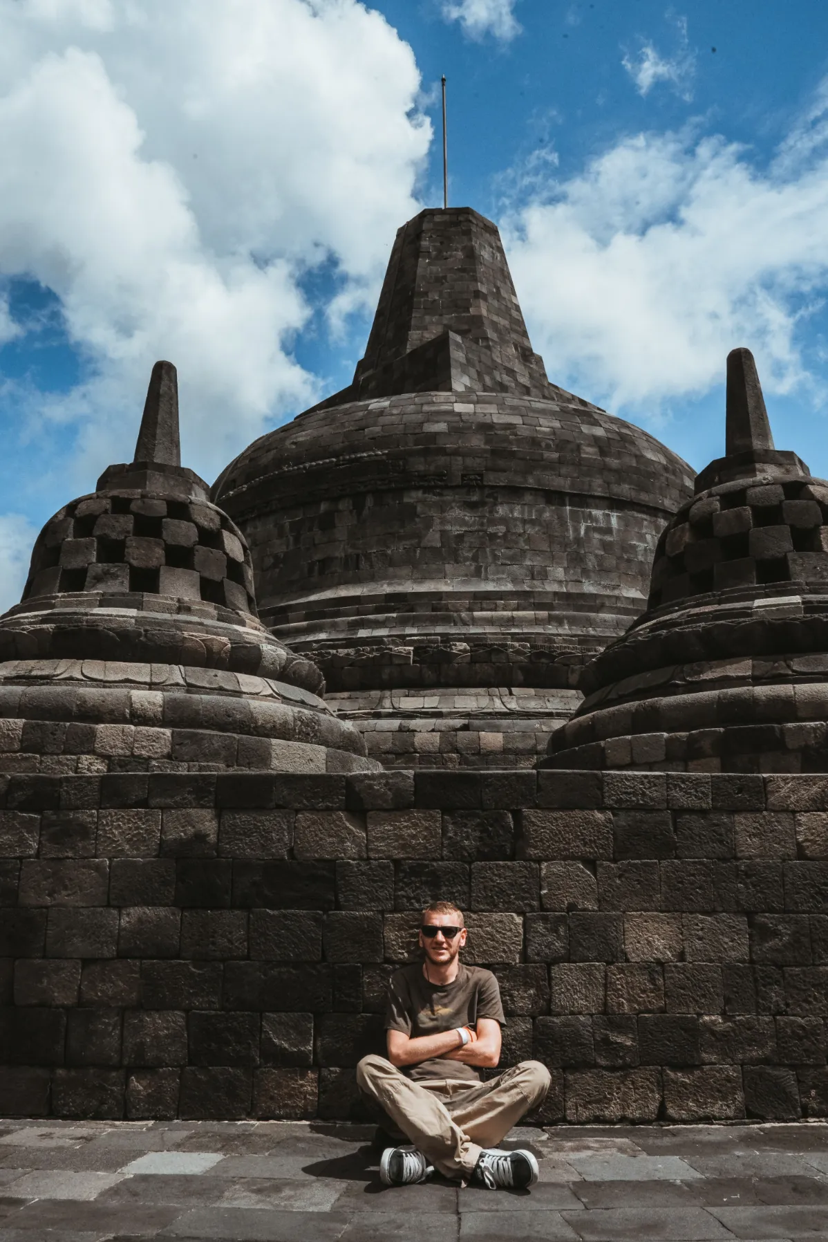 A person sits cross-legged in front of a large ancient temple, surrounded by dramatic clouds and a bright blue sky.