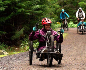 A person with a red helmet rides a handcycle energetically on a forest trail, followed by others. The scene is lively and surrounded by lush greenery.