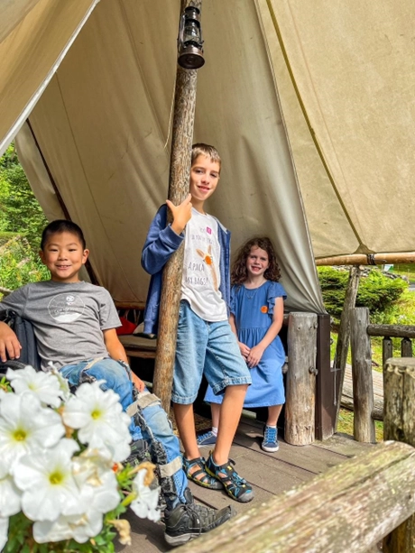 Three children are seated under a canvas tent, surrounded by greenery and white flowers, enjoying a sunny day outdoors.
