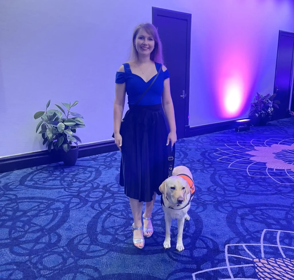 Woman in a blue top and black skirt with a guide dog wearing an orange vest.