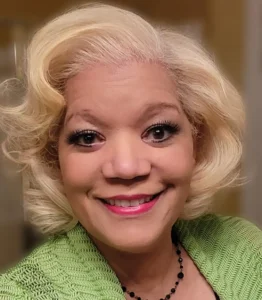 A woman with stylish, wavy blonde hair wearing a green cardigan and a beaded necklace, set against a softly blurred background.
