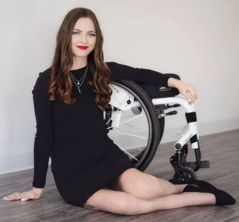 A woman in a black dress sits on the floor beside a wheelchair. She smiles confidently, with long brown hair draped over her shoulders, exuding empowerment.