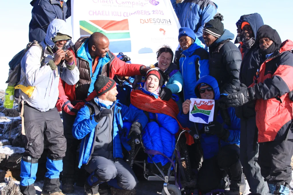 A celebratory group of climbers poses atop a mountain, holding a South African flag beside a poster for the Chaeli Kili campaign.