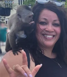 A woman smiling with a small monkey on her shoulder, outside. She gestures playfully with her hand. The scene feels joyful and casual.