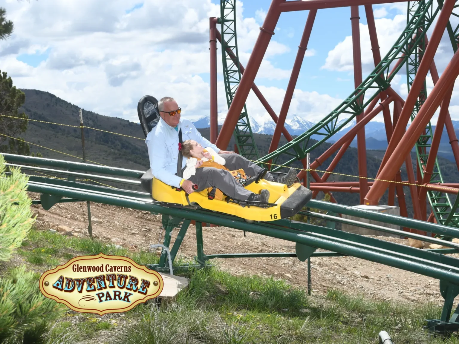 Two people ride a mountain coaster at Glenwood Caverns Adventure Park, with dramatic clouds and mountain scenery in the background.