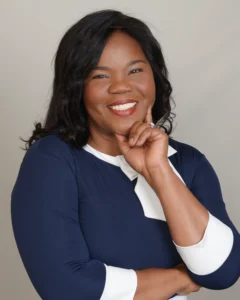 A woman in a navy blue dress with white accents poses thoughtfully, arms crossed, against a plain background.