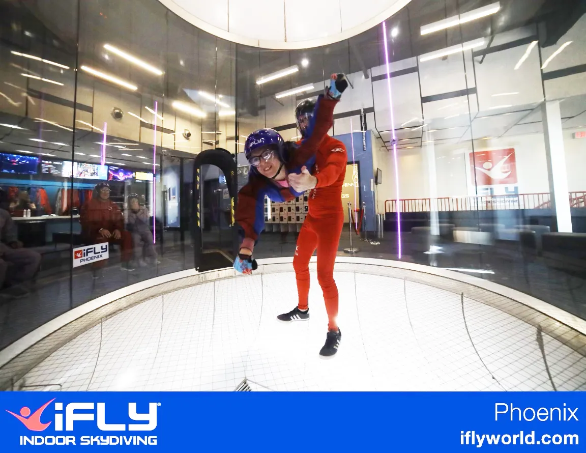 Person in red skydiving suit performing an indoor skydiving maneuver inside a large, round wind tunnel.