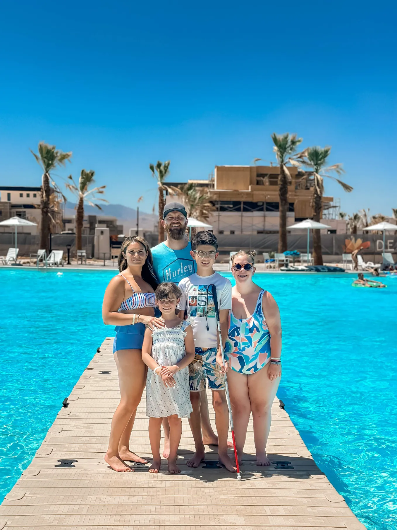A family of five poses on a wooden dock by a bright blue swimming pool, surrounded by palm trees under a clear sky.