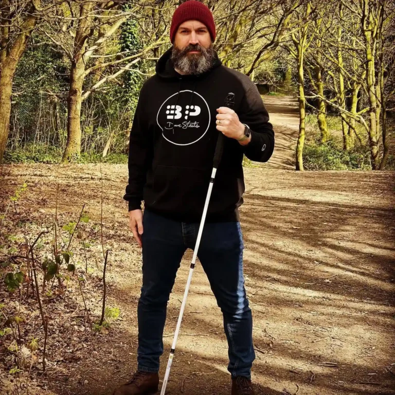 Man in black hoodie with white cane, outdoors in a forest.