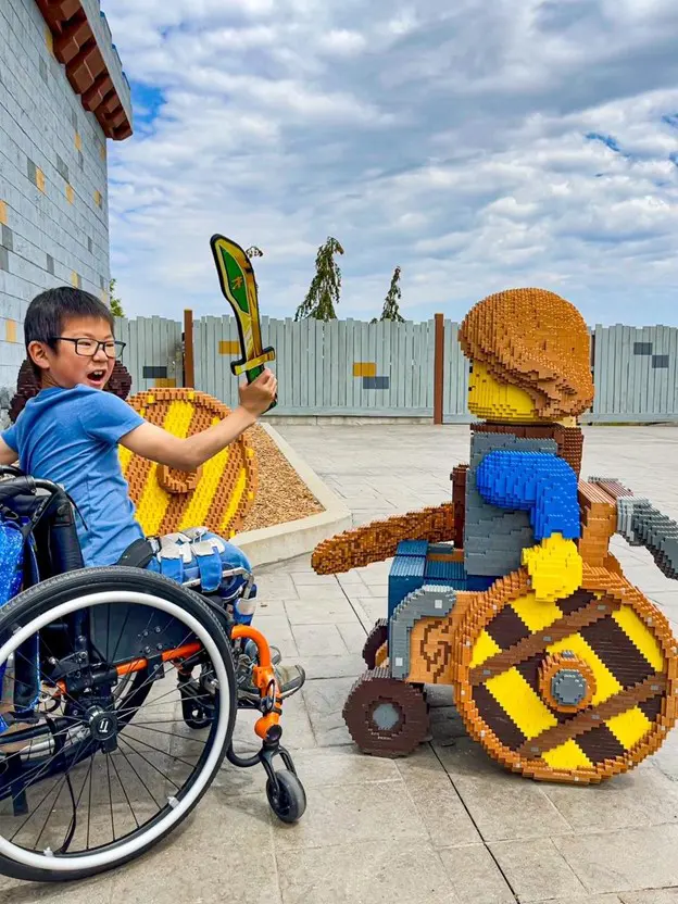 Two children, one in a wheelchair, admire a whimsical outdoor scene featuring large, colorful statues of animals and nature.