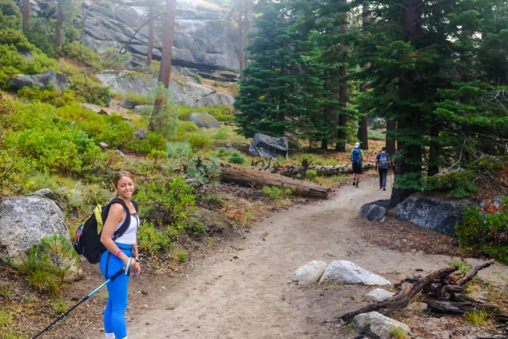 A serene hiking trail surrounded by tall trees, boulders, and lush greenery, with two hikers walking in the background.