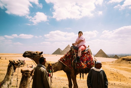 A person rides a decorated camel in front of the Pyramids of Giza, surrounded by two men in traditional attire under a bright sky.
