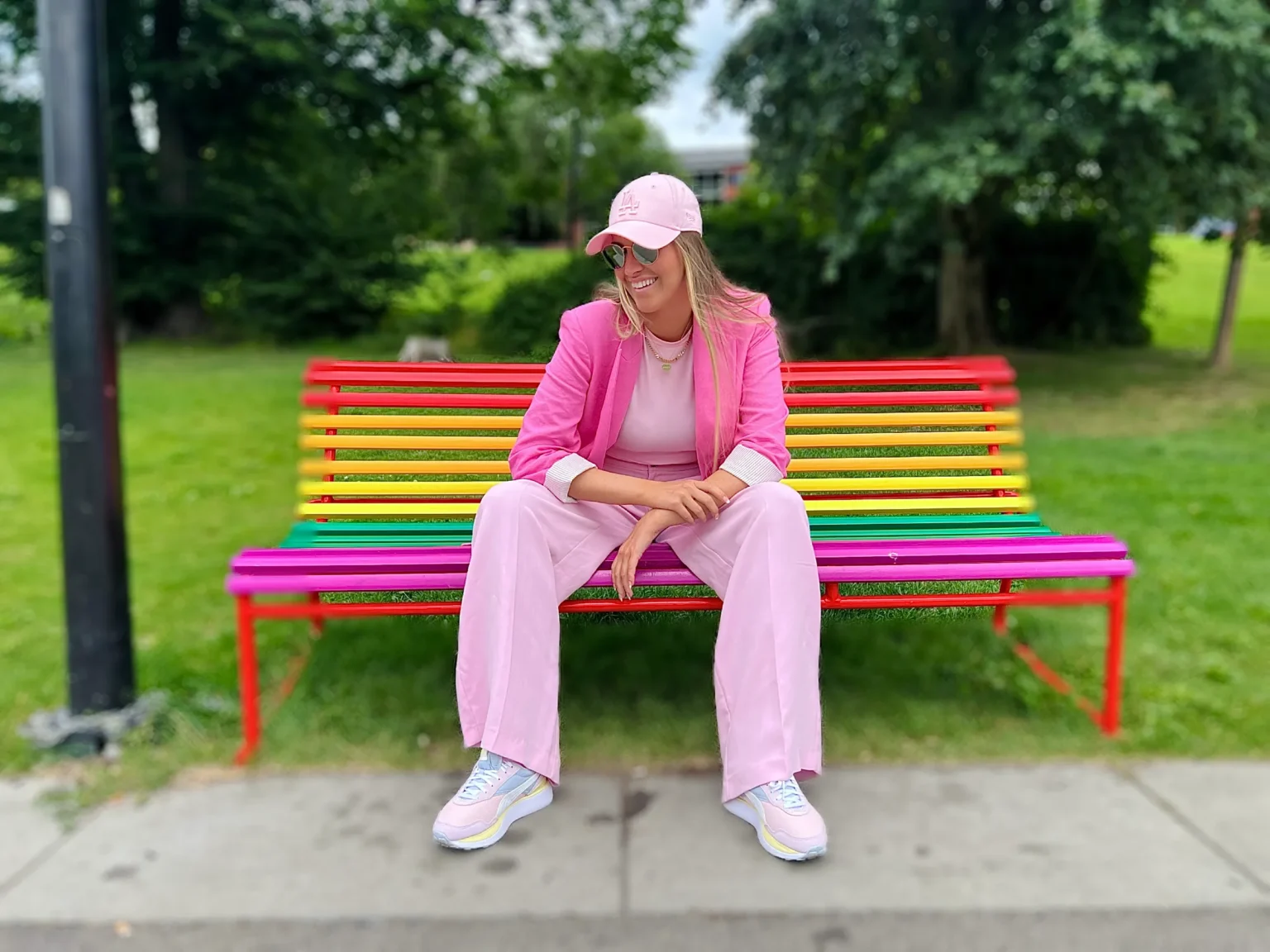 A person dressed in a pink suit sits on a colorful striped bench in a park, surrounded by greenery.