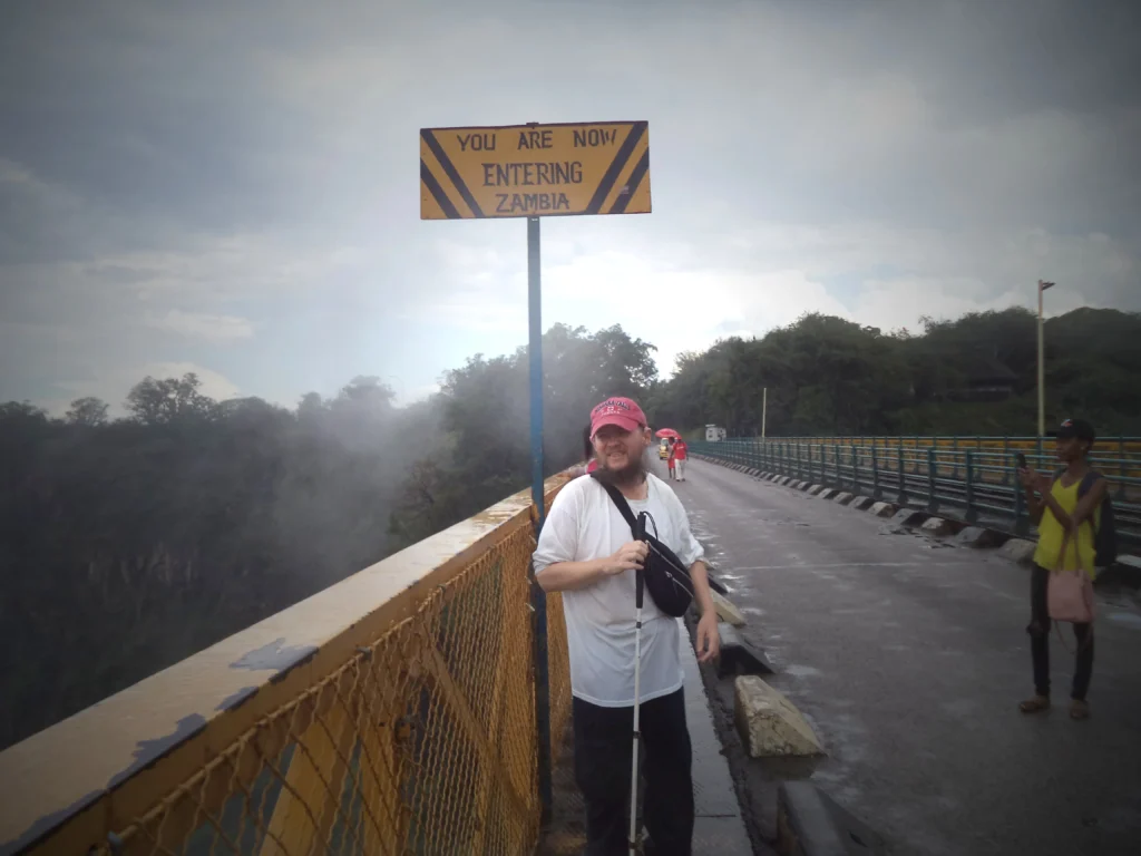 A person stands on a bridge under a sign reading "You Are Now Entering Zambia," with misty trees and another individual in the background.