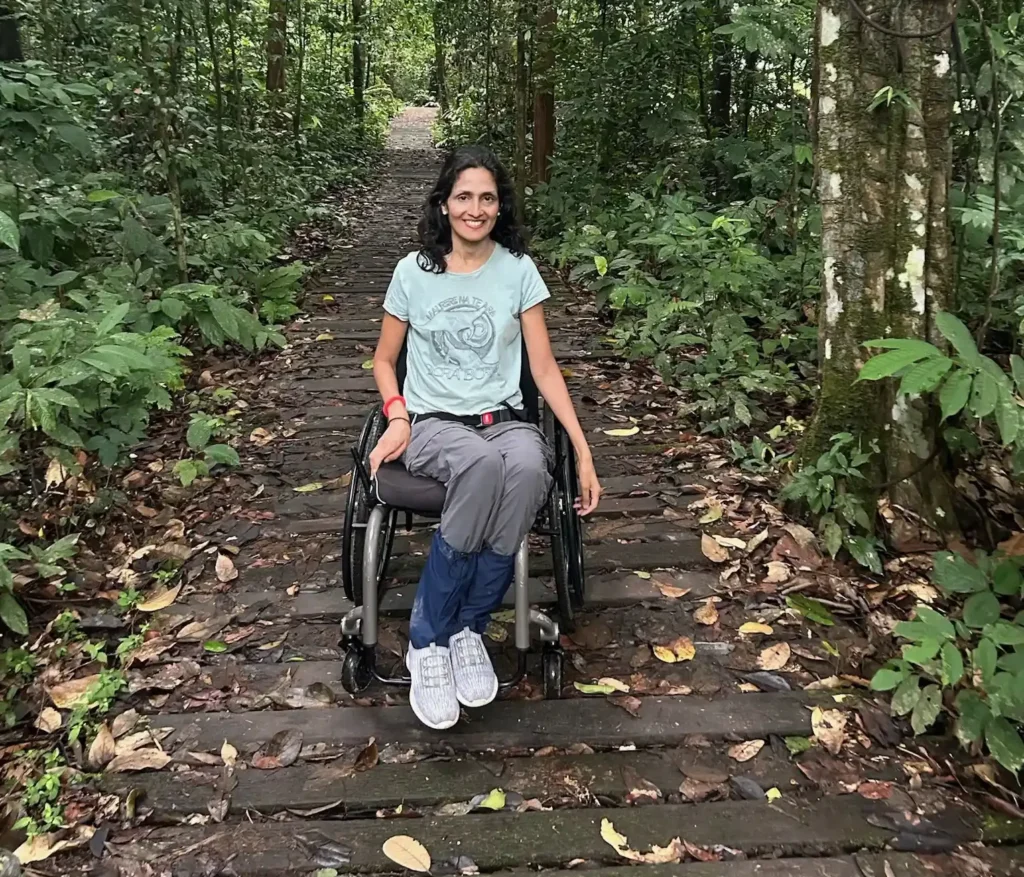 A person sitting in a wheelchair on a wooden path surrounded by lush green forest vegetation and scattered leaves.