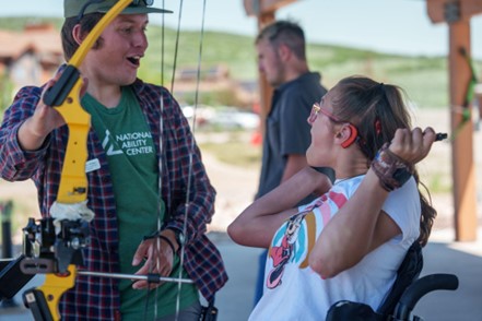 A person in a wheelchair prepares to shoot an arrow while assisted by another individual at an outdoor archery range.