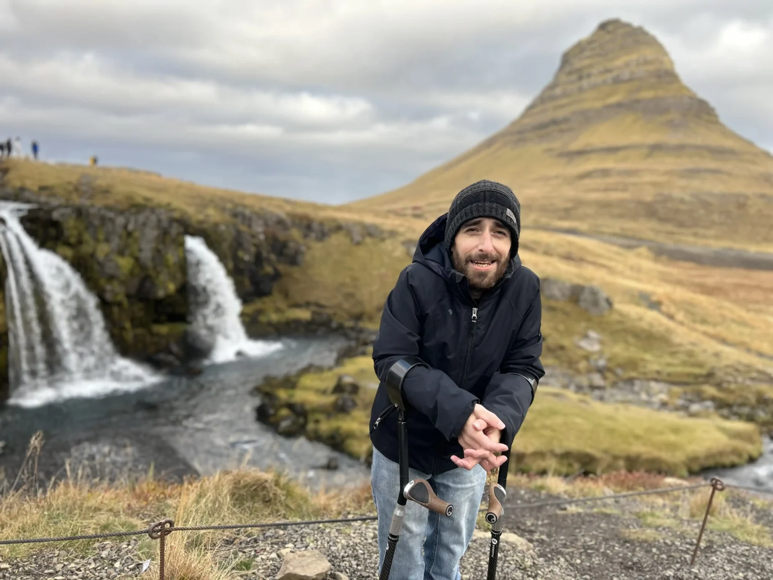 A person wearing a black jacket stands near scenic waterfalls, with a mountain and grassy landscape in the background.