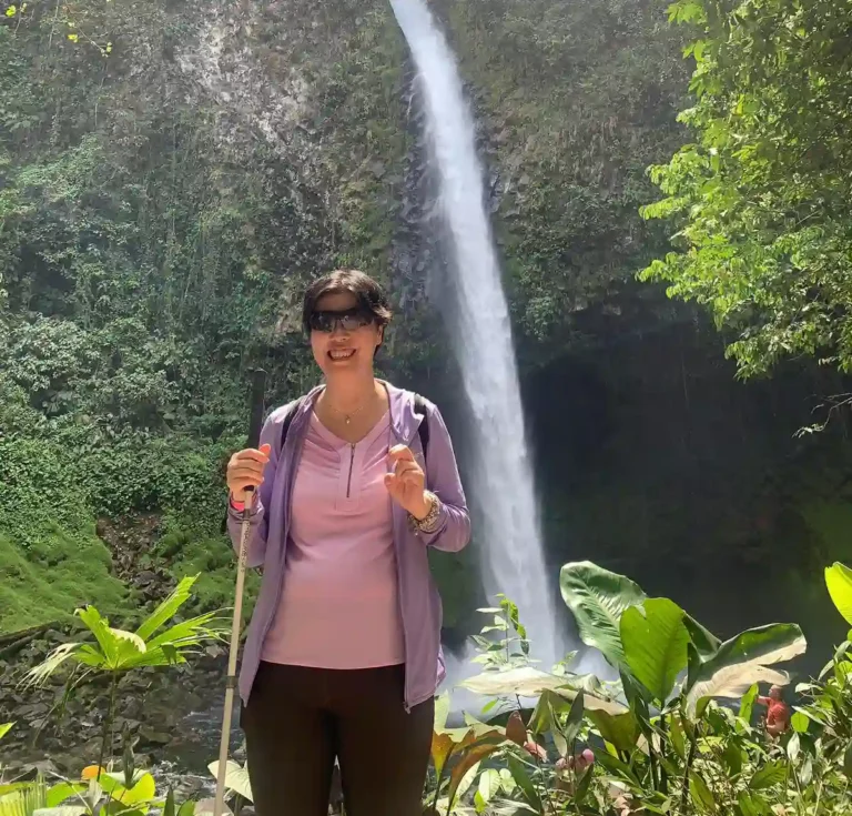 A person stands near a lush, green area with vibrant plants, in front of a tall waterfall cascading down a rocky cliff behind them.