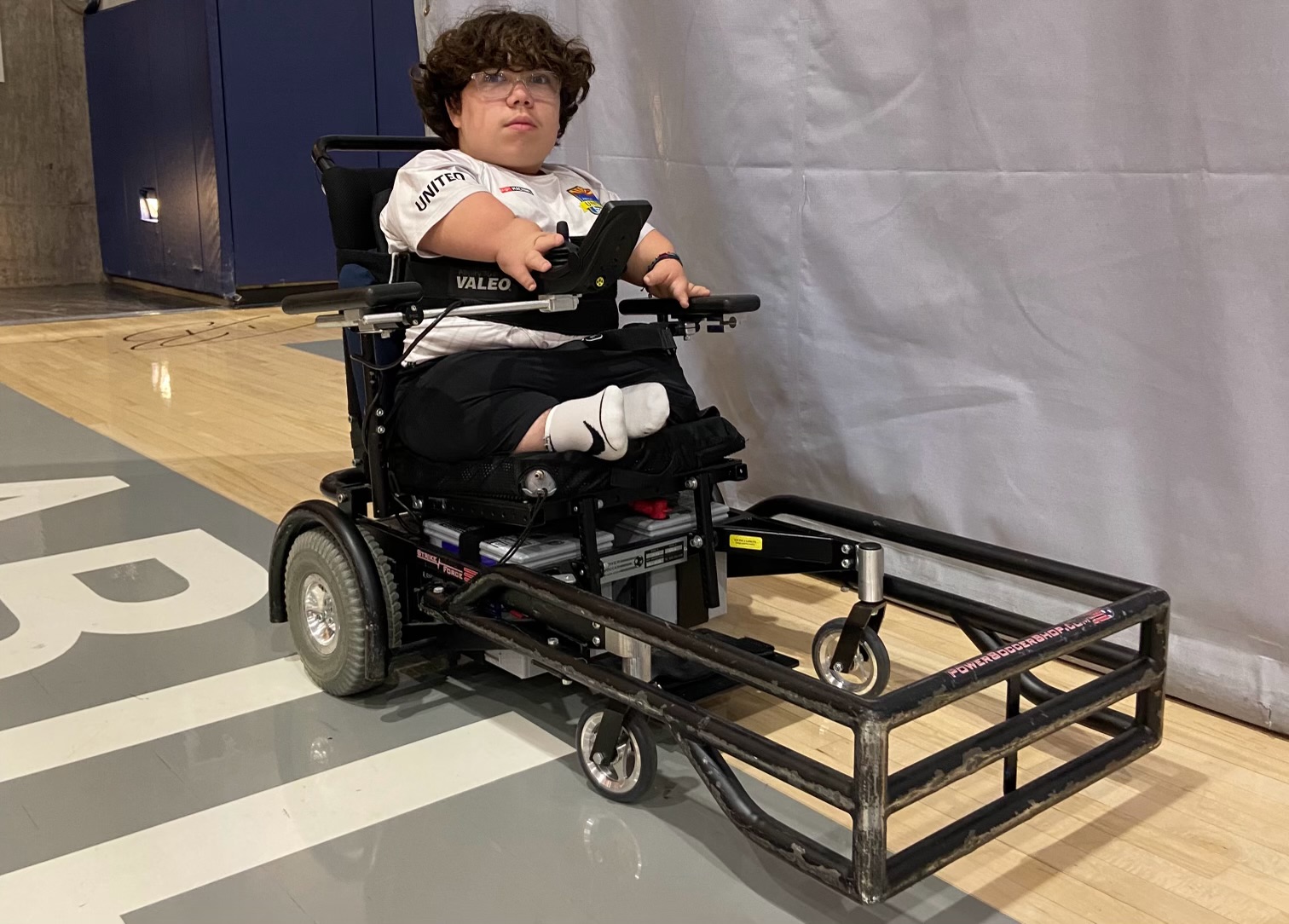 A person sits in a powered wheelchair, equipped with a support frame, on a gymnasium floor marked with white lines.