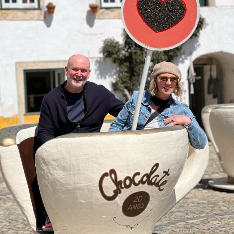 A couple sits inside a giant chocolate cup with a heart-shaped sign, enjoying a sunny day in a quaint outdoor setting.