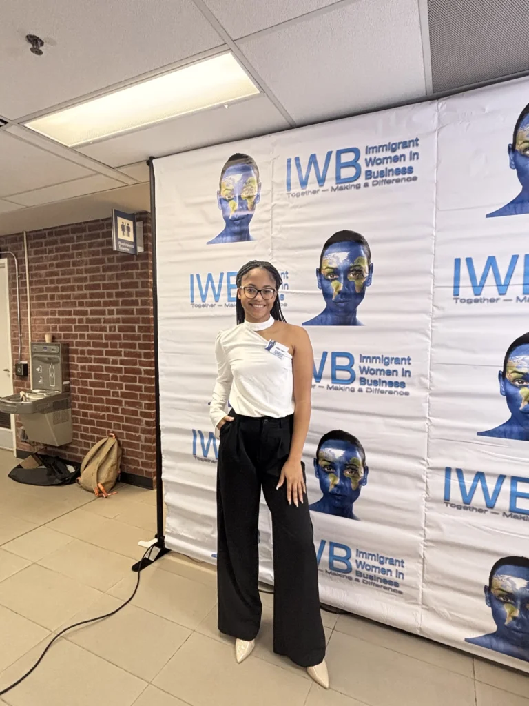 Young woman in stylish black pants and a white top, posing in front of a banner promoting Immigrant Women in Business.