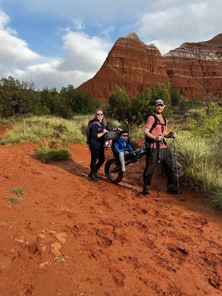 Two hikers pause on a reddish dirt trail surrounded by lush greenery and towering rock formations under a partly cloudy sky.