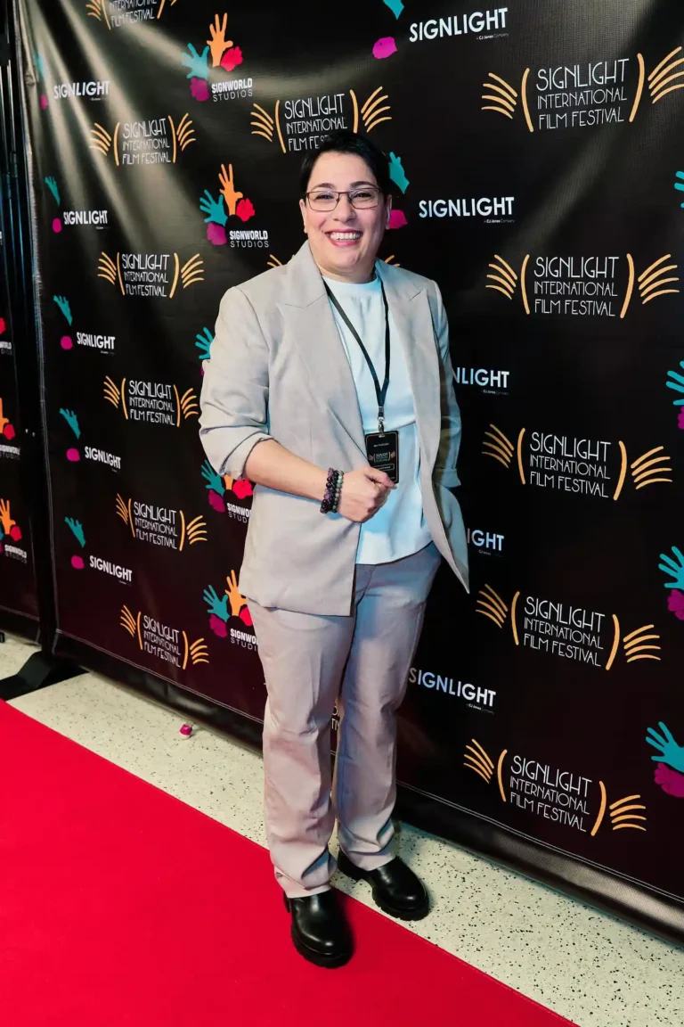 Person in a light gray suit smiling on a red carpet. Background features a black step-and-repeat banner with colorful logos and text.
