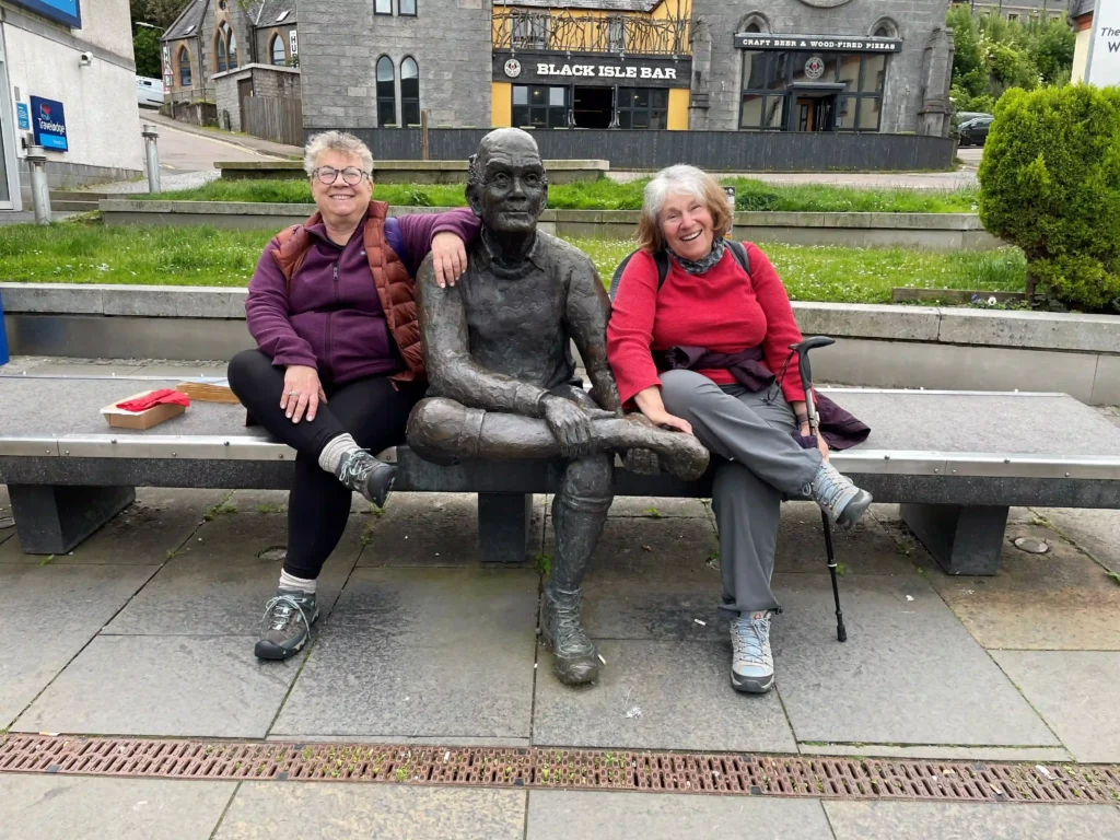 Two women sit on a bench beside a life-sized bronze statue, with a bar and greenery visible in the background.