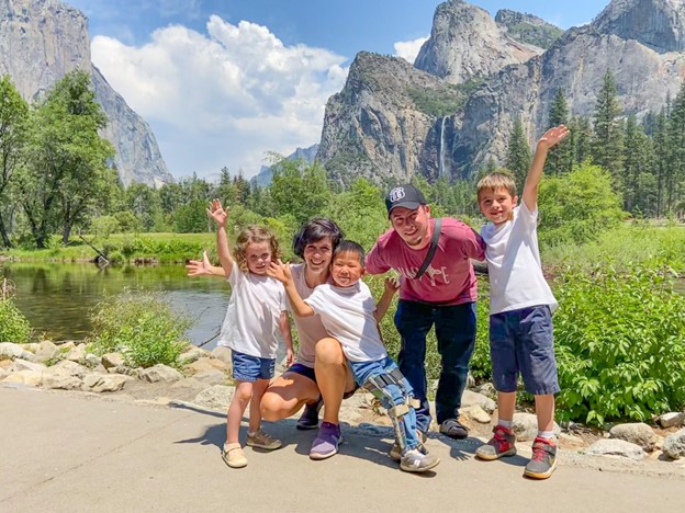 A group of four children and one adult pose playfully by a serene river, surrounded by towering mountains and lush greenery.