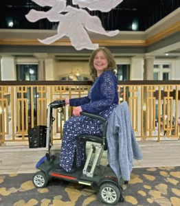A woman in a blue dress sits on a mobility scooter, smiling, on a balcony with decorative lights above.