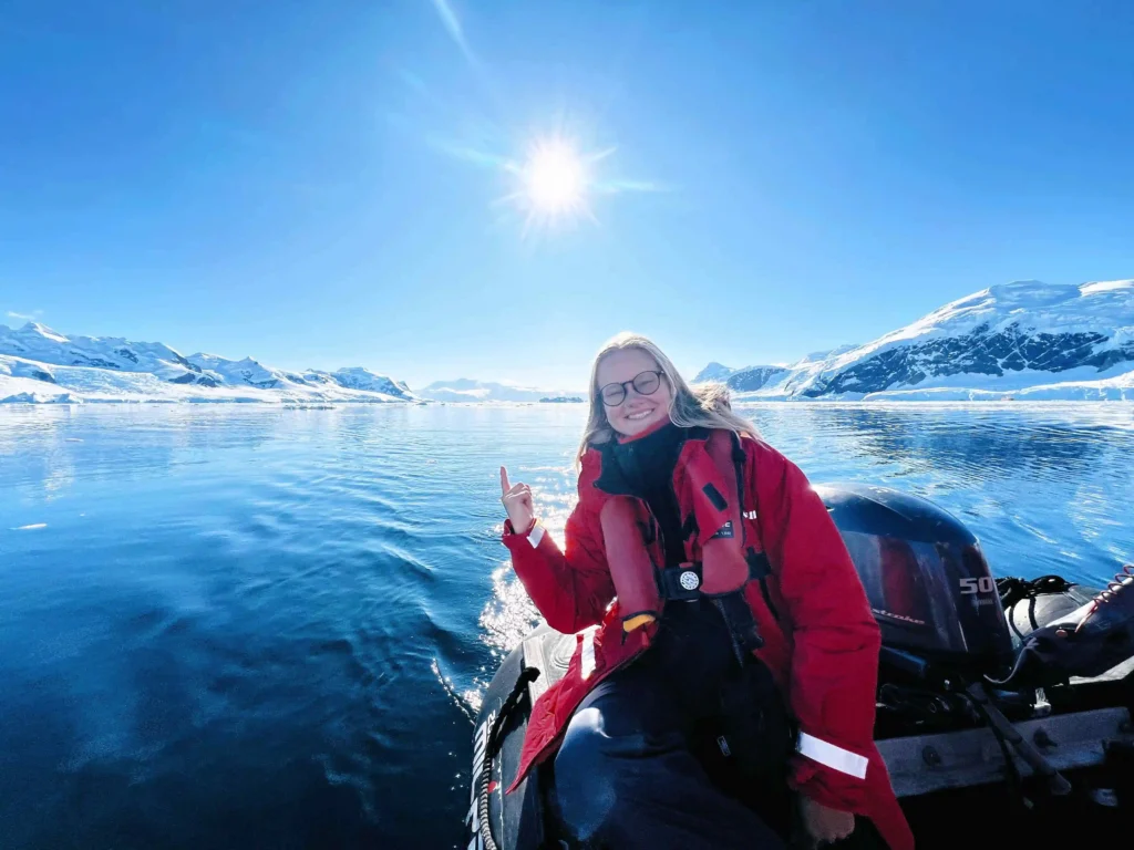 Woman in red life vest on a boat, pointing at snow-capped mountains, sunny Antarctic landscape.